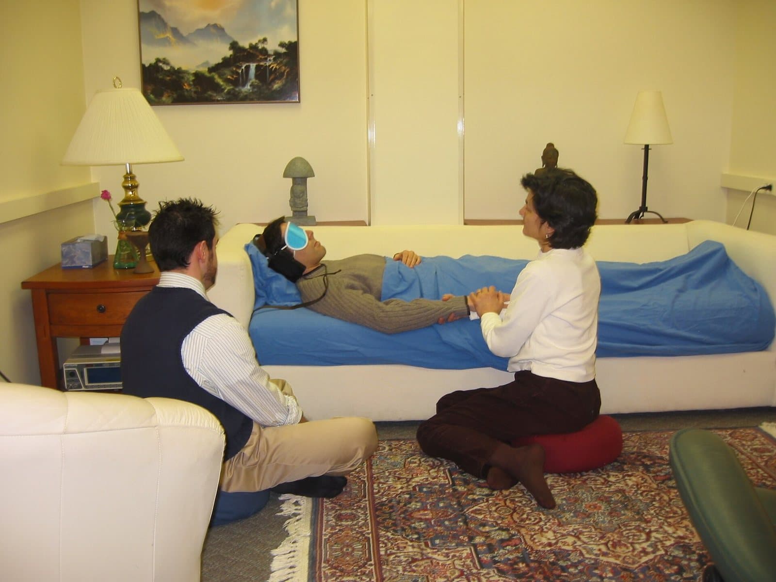 A participant wearing eyeshades lies on a couch under a blue blanket in a plainly furnished room while two session monitors sit beside them.
