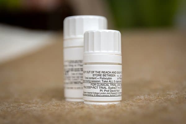 Two small white pharmacy bottles on a pale surface, the front one labeled for clinical trial use with psilocybin dosing instructions.